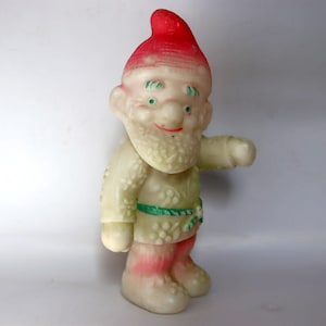 May include: A vintage gnome figurine with a red hat, white beard, and green eyes. The gnome wears a cream-colored outfit with a green belt and red-striped boots. The figurine is standing and has one arm outstretched.