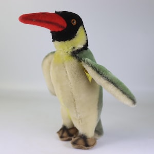 Puede incluir: Un juguete de peluche de tucán vintage. El pájaro de peluche tiene un pico rojo, cabeza negra, pecho amarillo y alas verdes. El juguete está de pie con las alas ligeramente extendidas. Los pies del pájaro son marrones.