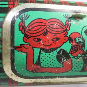 May include: Vintage metal lunchbox with a red-haired girl illustration on a green background. The girl is wearing a green polka-dot dress. The lunchbox has a red, black, and green plaid pattern on the sides and a cream-colored handle.