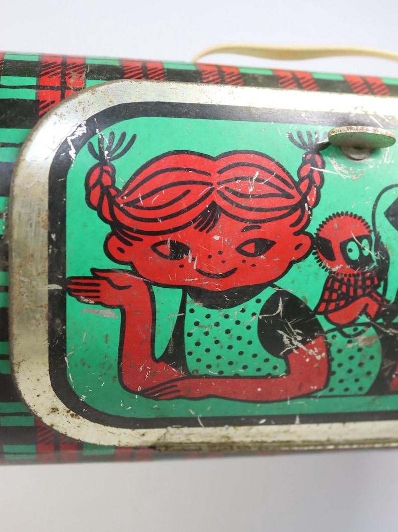May include: Vintage metal lunchbox with a red-haired girl illustration on a green background. The girl is wearing a green polka-dot dress. The lunchbox has a red, black, and green plaid pattern on the sides and a cream-colored handle.
