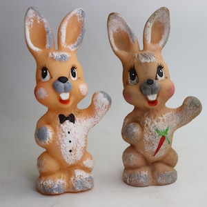 May include: Two vintage Easter bunny figurines. One bunny is wearing a black bow tie and a tuxedo-style shirt. The other bunny has a carrot design on its chest. Both bunnies have rosy cheeks, gray accents, and are in a waving pose.