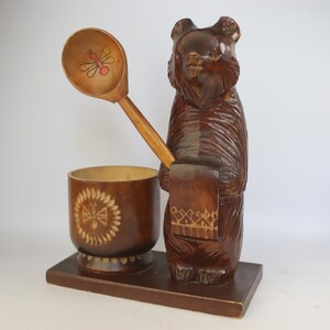 May include: A wooden bear figurine holding a spoon and a small container. The spoon has a bee design. The bear, spoon, and container are brown. The container has a decorative design. The items are on a rectangular base.