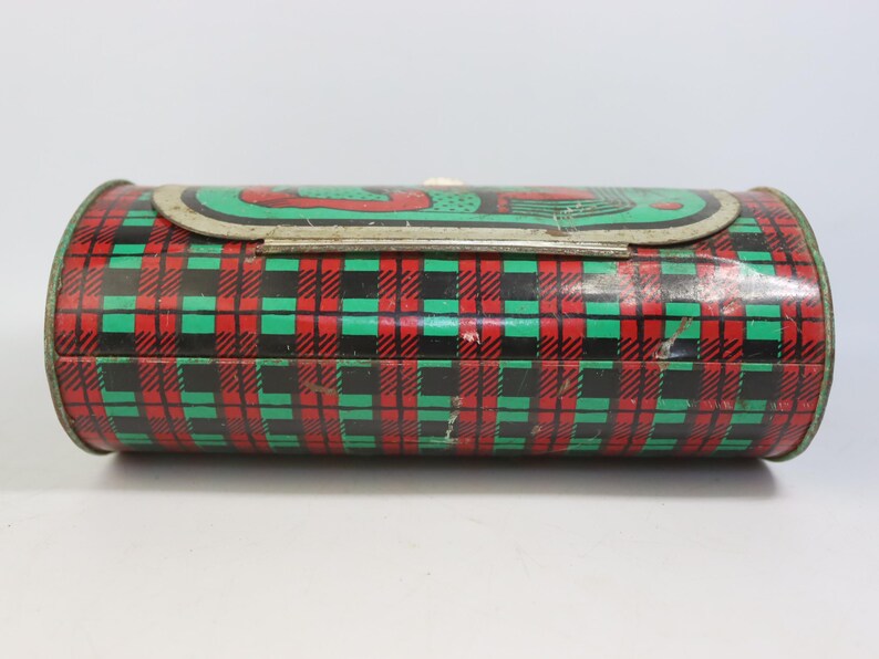 May include: A vintage, cylindrical tin box with a plaid pattern in red, green, and black. The lid features a green and red design. The metal box has a clasp closure and is likely used for storage or as a decorative item.
