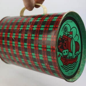 May include: Vintage cylindrical tin with a red, green, and black plaid pattern. One end features a green oval with a red sailboat illustration and the text "MADE IN USSR". The tin has a cream-colored handle.