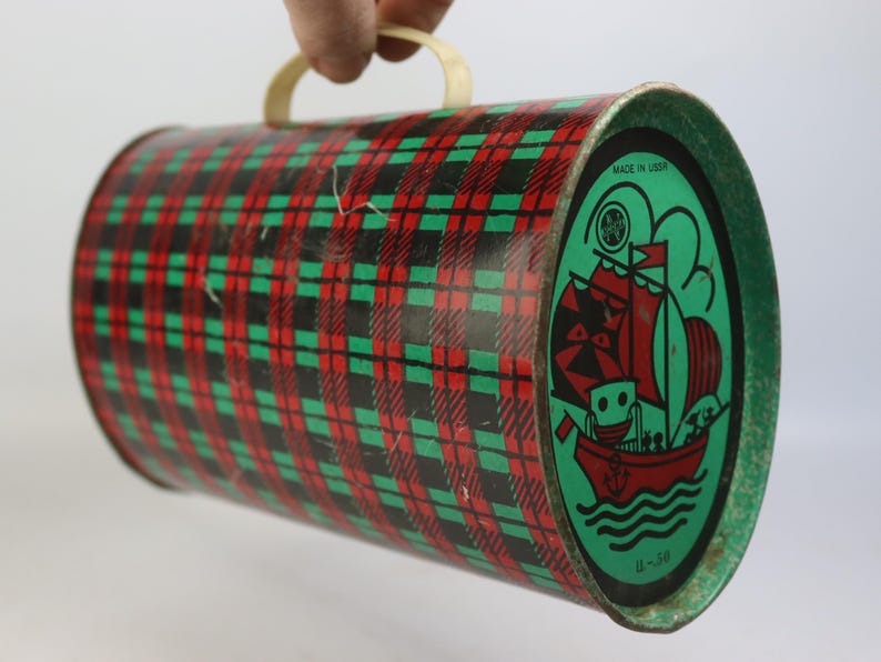 May include: Vintage cylindrical tin with a red, green, and black plaid pattern. One end features a green oval with a red sailboat illustration and the text "MADE IN USSR". The tin has a cream-colored handle.