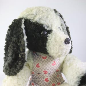 May include: A soft toy dog with black and white fur, wearing a grey apron patterned with red strawberries. The toy has floppy ears and a black nose, set against a white background.