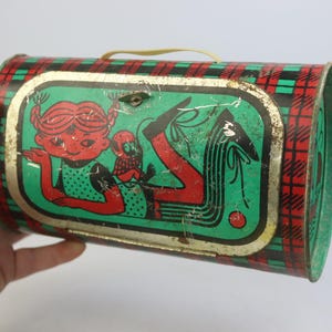 May include: Vintage metal lunchbox with a red, green, and black plaid pattern. The front features a cartoon illustration of a girl with red hair and a small creature. The lunchbox has a yellow handle and a metal clasp.