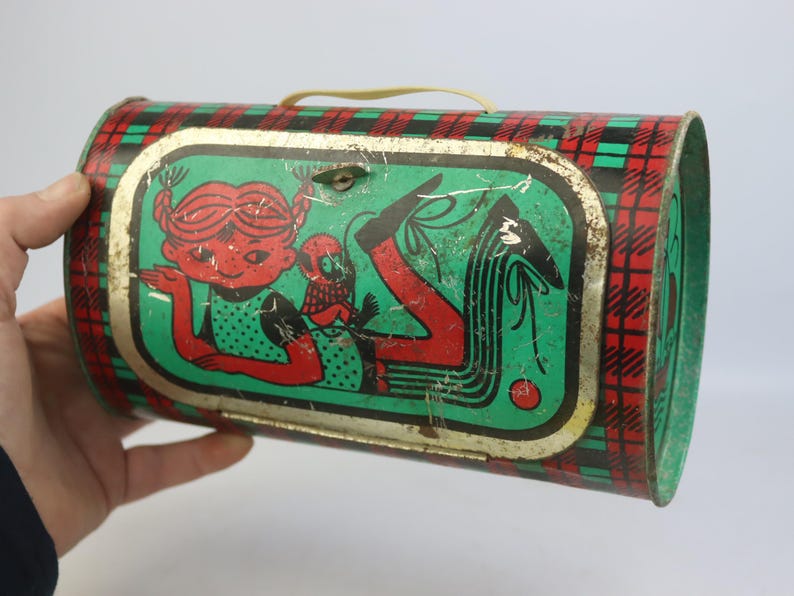 May include: Vintage metal lunchbox with a red, green, and black plaid pattern. The front features a cartoon illustration of a girl with red hair and a small creature. The lunchbox has a yellow handle and a metal clasp.
