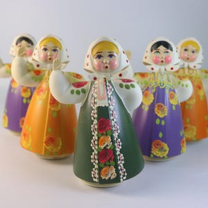 May include: A group of five Russian nesting dolls, each with a unique design. The dolls wear colorful dresses in orange, purple, and green, adorned with floral patterns. They have white headscarves and painted faces, creating a charming folk art aesthetic.