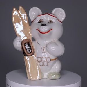 May include: A white ceramic bear figurine holding skis. The bear has a red headband, eyelashes, and a red smile. It wears a gold necklace with a number one pendant and the Olympic rings. The skis are light brown.