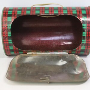 May include: Vintage metal lunchbox with a red, green, and black plaid pattern. The lunchbox has a brown interior and a hinged lid. A small, tan handle is attached to the top. The lunchbox is open, revealing the interior.