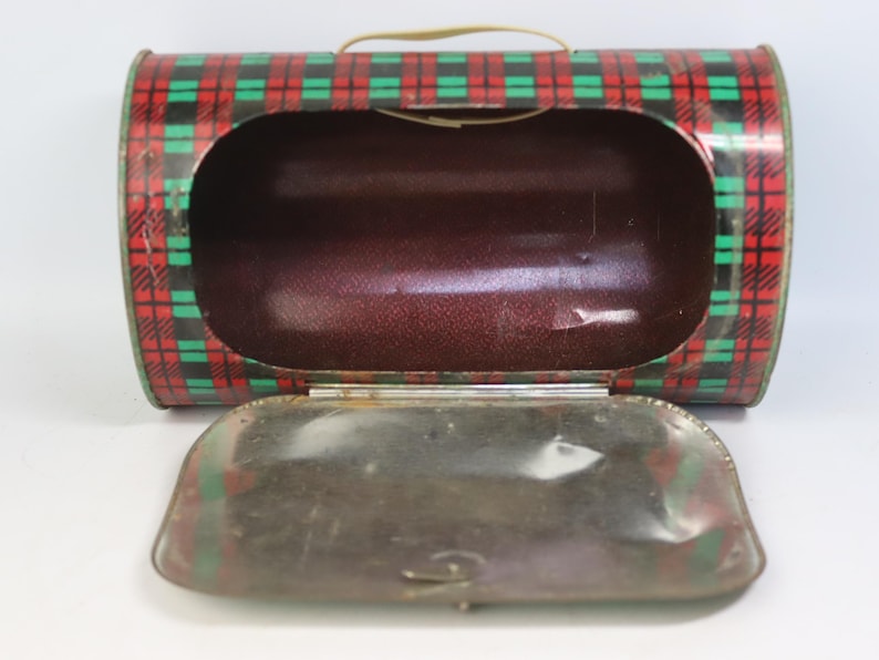 May include: Vintage metal lunchbox with a red, green, and black plaid pattern. The lunchbox has a brown interior and a hinged lid. A small, tan handle is attached to the top. The lunchbox is open, revealing the interior.