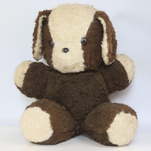 Pode incluir: Um cão de peluche com pelo castanho escuro e creme. O brinquedo apresenta uma mancha creme na cabeça e no rosto, orelhas castanho escuro, um corpo castanho escuro e patas creme. Tem pequenos olhos e nariz de plástico preto, redondos e brilhantes.