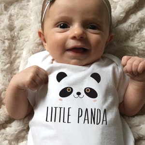 May include: A white baby onesie with a black and white panda face graphic and the text "LITTLE PANDA". The baby is wearing a white headband with a silver bow.