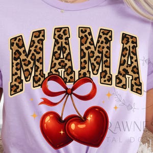 Puede incluir: Una camiseta de color lila claro con la palabra "MAMA" en un diseño de estampado de leopardo. Debajo del texto hay dos cerezas rojas brillantes en forma de corazón atadas con un lazo rojo. La camiseta es de cuello redondo.