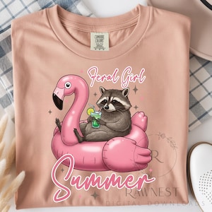 May include: A light pink t-shirt featuring a raccoon relaxing on a pink flamingo float with a drink. The words "Feral Girl Summer" are written in a script font. The shirt is made of Comfort Colors material.