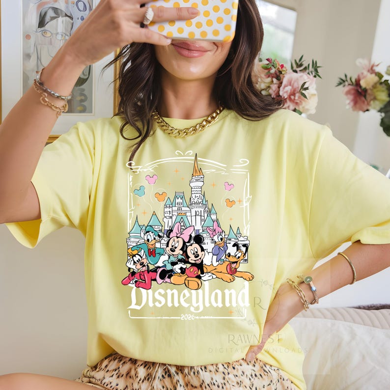 Peut inclure: Un t-shirt jaune p&acirc;le avec une impression graphique repr&eacute;sentant un ch&acirc;teau et divers personnages de dessins anim&eacute;s, dont Mickey, Minnie, Donald, Daisy, Dingo et Pluto. Le texte "Disneyland" et "2026" est imprim&eacute; sous les personnages.