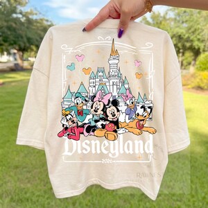 Peut inclure: Un t-shirt couleur cr&egrave;me avec une impression graphique repr&eacute;sentant un ch&acirc;teau et des personnages Disney dont Mickey, Minnie, Donald, Daisy, Dingo et Pluto. Le texte Disneyland et 2026 est imprim&eacute; sous les personnages.
