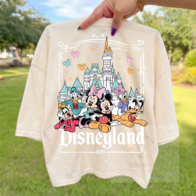 Peut inclure: Un t-shirt couleur cr&egrave;me avec une impression graphique repr&eacute;sentant un ch&acirc;teau et des personnages Disney dont Mickey, Minnie, Donald, Daisy, Dingo et Pluto. Le texte Disneyland et 2026 est imprim&eacute; sous les personnages.