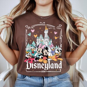 Peut inclure: Un t-shirt marron &agrave; manches courtes avec une impression graphique repr&eacute;sentant un ch&acirc;teau blanc, Mickey, Minnie, Donald, Daisy, Dingo et Pluto. Le texte Disneyland 2026 est imprim&eacute; sous les personnages avec des ballons flottants.