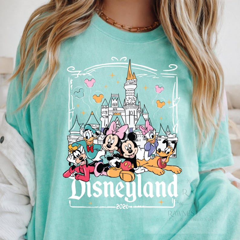 Peut inclure: Un t-shirt vert menthe avec une impression graphique repr&eacute;sentant un ch&acirc;teau et des personnages Disney dont Mickey, Minnie, Donald, Daisy, Dingo et Pluto. Le texte Disneyland 2026 est imprim&eacute; en blanc sous les personnages.