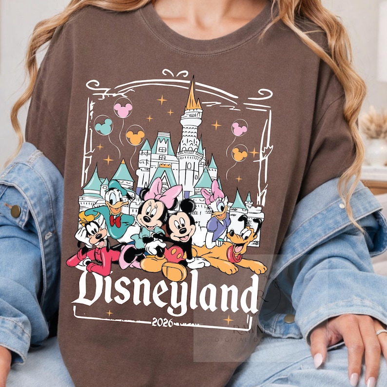 Peut inclure: Un sweat-shirt marron &agrave; col rond avec une impression graphique repr&eacute;sentant un ch&acirc;teau blanc, des ballons color&eacute;s et des personnages de dessins anim&eacute;s dont Mickey, Minnie, Donald, Daisy, Dingo et Pluto avec le texte Disneyland 2026.