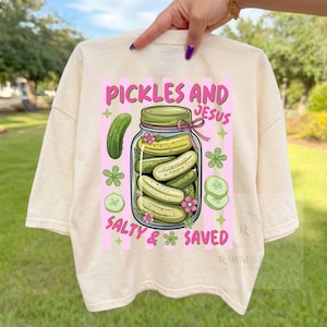 Peut inclure: T-shirt crème avec un motif graphique. Le motif comprend le texte "Pickles and Jesus", un bocal de cornichons, un seul cornichon et les mots "Salty & Saved".