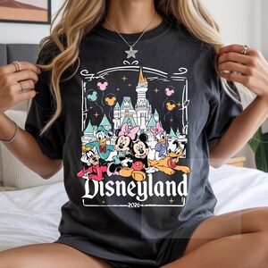 Peut inclure: Un t-shirt gris anthracite avec une impression graphique repr&eacute;sentant un ch&acirc;teau, Mickey, Minnie, Donald, Daisy, Dingo et Pluto. Le texte Disneyland et 2026 est imprim&eacute; sous les personnages, entour&eacute; d'un cadre d&eacute;coratif blanc.