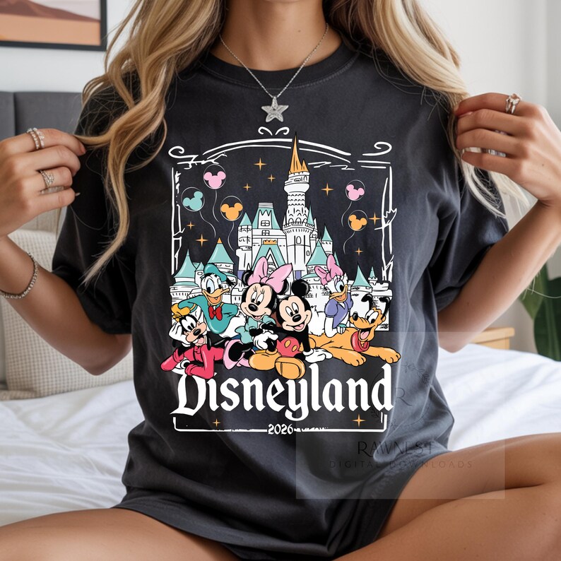 Peut inclure: Un t-shirt gris anthracite avec une impression graphique repr&eacute;sentant un ch&acirc;teau, Mickey, Minnie, Donald, Daisy, Dingo et Pluto. Le texte Disneyland et 2026 est imprim&eacute; sous les personnages, entour&eacute; d'un cadre d&eacute;coratif blanc.
