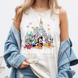 Peut inclure: Un d&eacute;bardeur sans manches de couleur cr&egrave;me avec une impression graphique de Mickey, Minnie, Donald, Daisy, Dingo et Pluto devant un ch&acirc;teau. Le texte Disneyland 2020 est imprim&eacute; sous les personnages dans une police de couleur blanche.