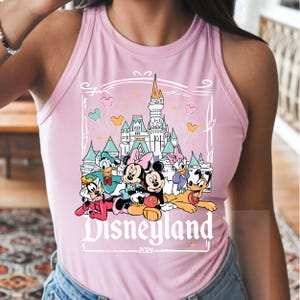 Peut inclure: Un d&eacute;bardeur rose clair sans manches avec une impression graphique repr&eacute;sentant un ch&acirc;teau, Mickey, Minnie, Donald, Daisy, Dingo et Pluto. Le texte Disneyland et 2026 est imprim&eacute; sous les personnages dans une police d&eacute;corative blanche.
