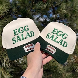 Può includere: Due cappellini da baseball con corona color crema e visiera verde scuro. Le parole "EGG SALAD" sono ricamate in verde sulla parte anteriore di ogni cappellino. I cappellini sono tenuti su uno sfondo di fogliame verde.