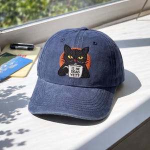 Puede incluir: Gorra de béisbol azul marino con visera curva. La gorra presenta un diseño bordado de un gato negro sosteniendo una taza con el texto "IS HE DEAD YET?" sobre un círculo naranja.