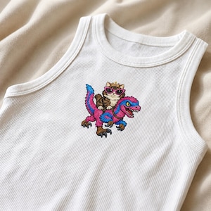 May include: White ribbed tank top featuring a colorful embroidered design. The design depicts a cat wearing sunglasses and a crown, riding a pink and blue dinosaur. The tank top has a classic, sleeveless design.
