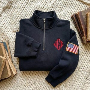 May include: A folded black sweatshirt with a quarter-zip collar. It features a red embroidered monogram on the chest and an American flag patch on the sleeve. The sweatshirt is displayed on a cream-colored, textured surface.