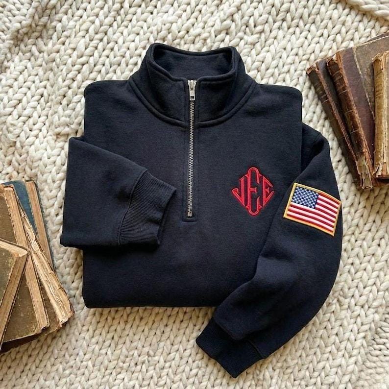 May include: A folded black sweatshirt with a quarter-zip collar. It features a red embroidered monogram on the chest and an American flag patch on the sleeve. The sweatshirt is displayed on a cream-colored, textured surface.