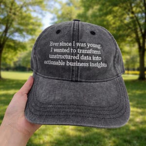 May include: A dark gray baseball cap with the text "Ever since I was young. I wanted to transform unstructured data into actionable business insights" embroidered in white. The cap is worn and has a curved brim.