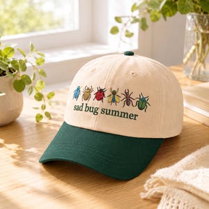 May include: A beige baseball cap with a dark green brim and button. The cap features embroidered colorful bugs and the text "sad bug summer" in green. The cap is made of a soft fabric.
