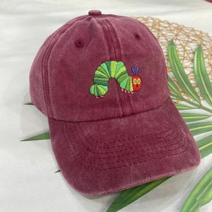 Puede incluir: Gorra de béisbol granate con visera curva. La gorra presenta un diseño bordado de una oruga verde y amarilla con detalles rojos. La gorra está hecha de un material suave y texturizado.