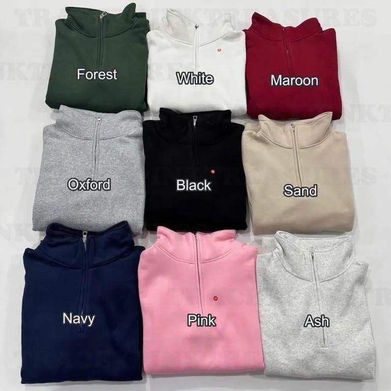 May include: A collection of nine quarter-zip sweatshirts in various colors, including forest green, white, and maroon. Each sweatshirt has a small embroidered detail and is labeled with its color name: Oxford, Black, Sand, Navy, Pink, and Ash.