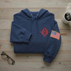 May include: Navy blue hoodie with a red embroidered monogram and an American flag patch on the sleeve. The hoodie has a drawstring hood and ribbed cuffs. A pair of glasses and a cup of coffee are also in the image.