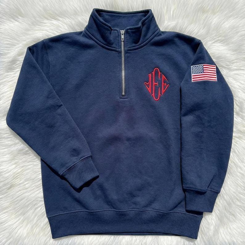May include: Navy blue quarter-zip sweatshirt with a red embroidered monogram and an American flag patch on the sleeve. The sweatshirt has a high collar and a silver zipper. The fabric appears to be a soft, comfortable material.