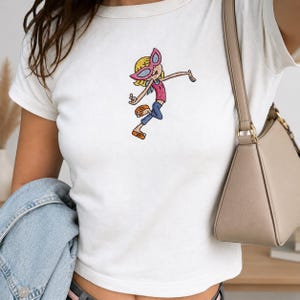 May include: A white short-sleeved t-shirt featuring a central embroidered graphic of a cartoon character with blonde hair, pink sunglasses, a pink top, blue pants, and orange shoes, depicted in a dynamic pose with one arm extended outward.