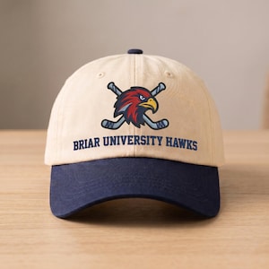 May include: A beige baseball cap with a navy blue brim. The cap features a logo of a red and yellow hawk with crossed hockey sticks and the text "BRIAR UNIVERSITY HAWKS" in navy blue.