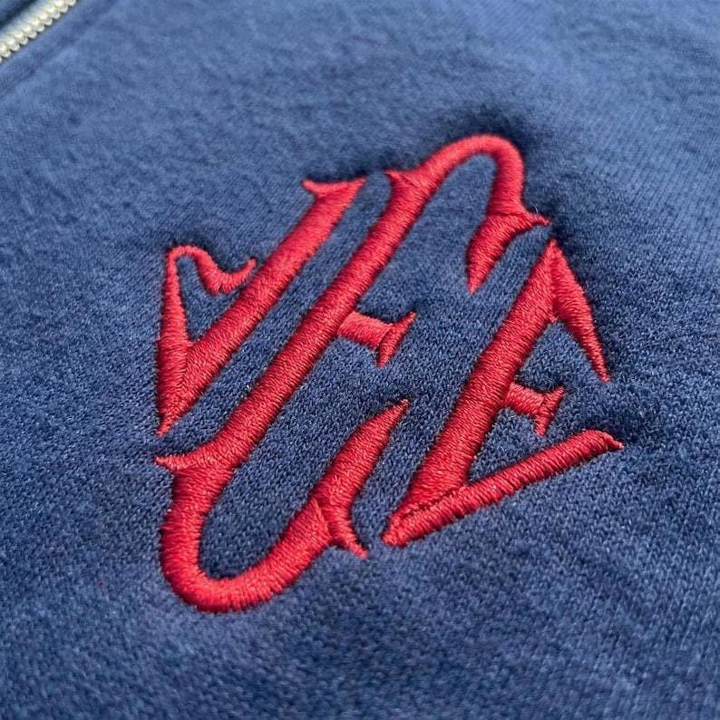 May include: Close-up of a navy blue zip-up hoodie with a red embroidered monogram. The monogram is a stylized, ornate design. The fabric has a soft, textured appearance.