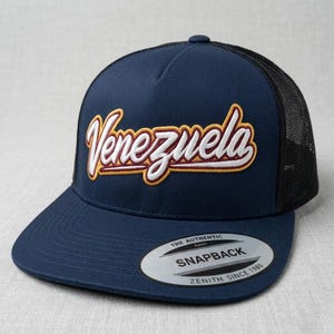 Może przedstawiać: Granatowa czapka typu snapback z czarną siatką z tyłu. Słowo "Venezuela" jest wyhaftowane kursywą z białymi, żółtymi i czerwonymi akcentami. Posiada zakrzywiony daszek i zapięcie na zatrzask.