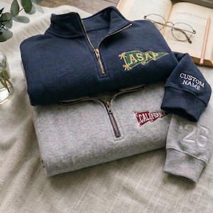 May include: Two folded sweatshirts, one navy blue and one gray, with quarter-zip closures. The navy sweatshirt has a gold embroidered "ASAP" design, and the gray one has a "CALIFORNIA" design. The navy sweatshirt cuff says "CUSTOM NAME", and the gray cuff has the number "26".