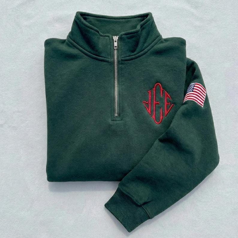 May include: Dark green quarter-zip sweatshirt with a red monogram and an American flag patch on the sleeve. The sweatshirt has a ribbed collar and cuffs, and a silver zipper. The monogram is in a diamond shape.