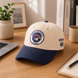 May include: A beige baseball cap with a navy blue brim. The cap features a circular patch with a hawk logo and the words "Briar University Hawks." The name "Graham" and the number "44" are on the side.