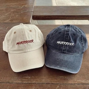 May include: Two baseball caps, one beige and one dark blue, are displayed on a wooden surface. Both caps have the word "MURRRDER" embroidered in a contrasting color. The beige cap has brown embroidery, while the dark blue cap has white embroidery.
